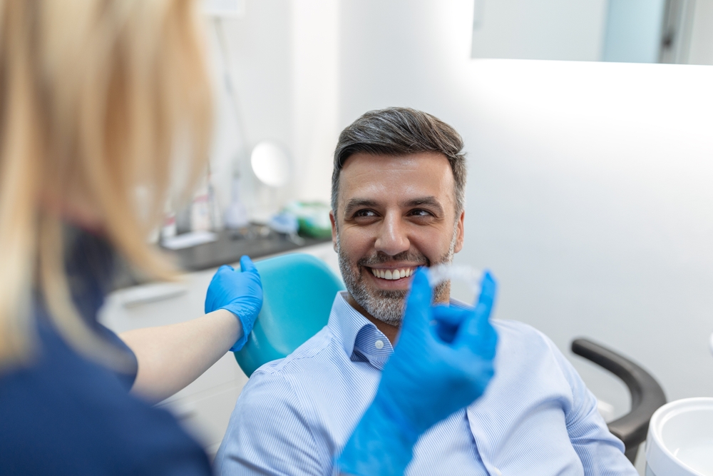 A dental professional showing an Invisalign clear aligner to a smiling male patient seated in a dental chair – Invisalign