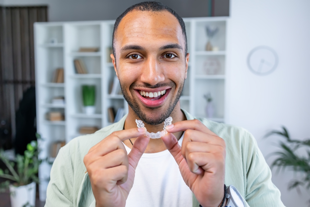 Smiling Man Holding Clear Aligner at Home – Dentist Orting WA A cheerful man stands indoors holding a clear aligner with both hands, showing his straight smile and satisfaction with orthodontic care – Dentist Orting WA