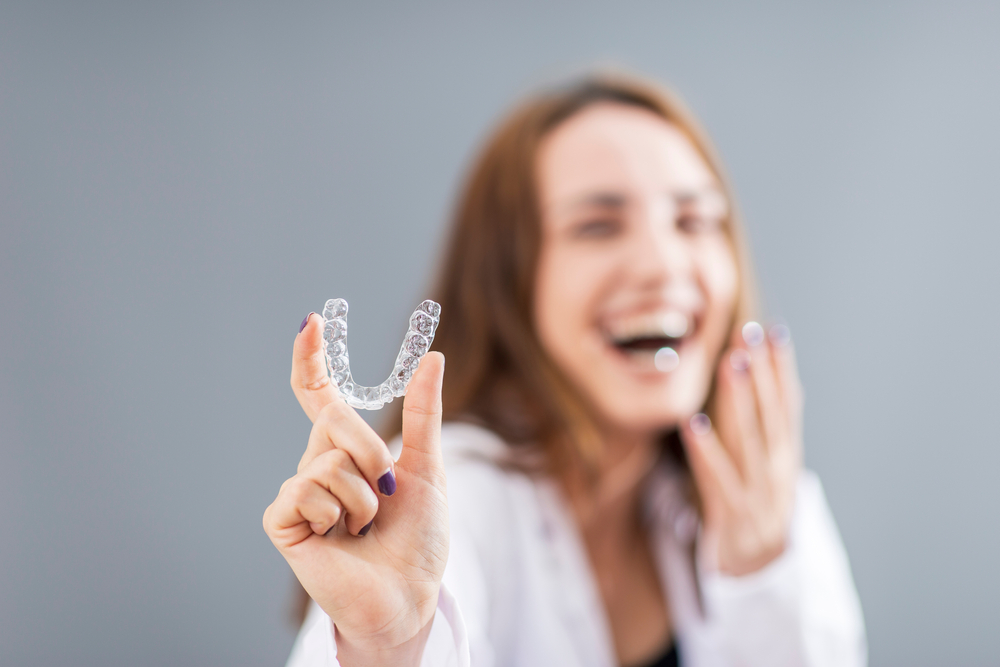 A happy woman laughing and holding up a clear Invisalign aligner, highlighting confidence and satisfaction with orthodontic treatment – Invisalign