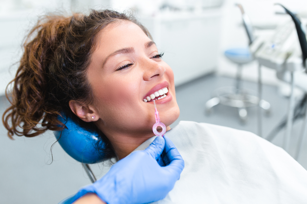 Woman Getting Tooth Shade Matched at Dental Office – Dental Crown A smiling woman reclines in a dental chair as a dentist matches her tooth color using a shade guide before placing a dental crown – Dental Crown