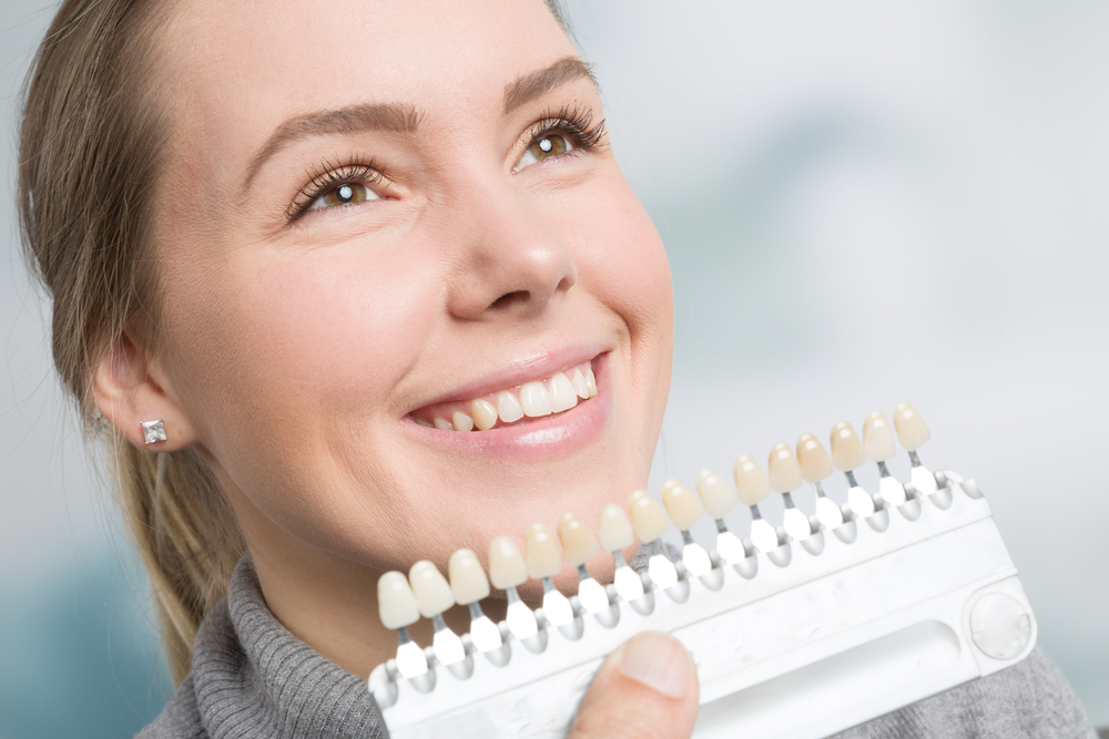 A woman smiles while holding a dental shade guide near her mouth during a cosmetic dentistry consultation, evaluating the best tooth color match – Dentist Spanaway WA