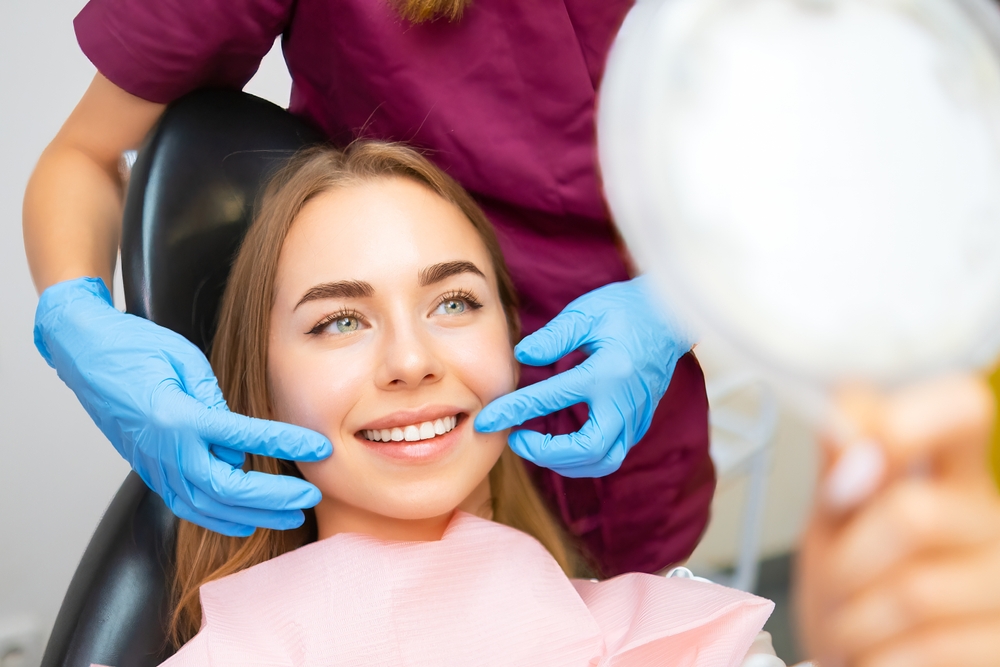 A young woman smiling while holding a mirror in a dental chair, as a dentist with blue gloves gently examines her mouth – Experiencing an Emergency?