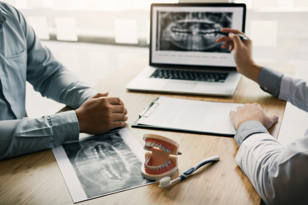 Dentist Reviewing Panoramic X-Ray with Patient – Dental X-Rays A dentist and patient reviewing a panoramic dental X-ray on a laptop during a consultation, with a model of teeth and toothbrush on the table – Dental