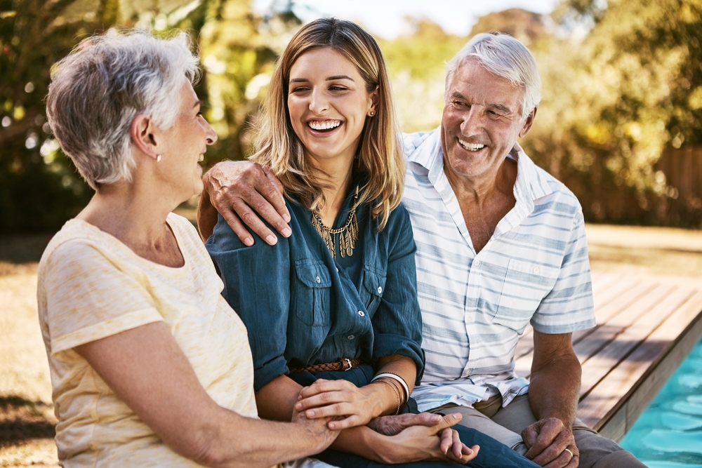 A cheerful middle-aged couple and young woman sitting by a pool, sharing a joyful moment together, representing the importance of oral health even in emergencies – Experiencing an Emergency?