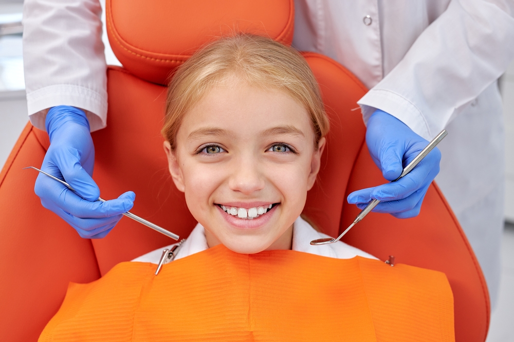 Smiling Young Girl During Dental Checkup – Dental Crown A young girl sitting in an orange dental chair, smiling confidently while a dentist with blue gloves prepares for her dental crown procedure – Dental Crown