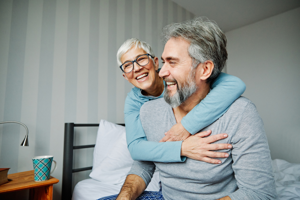 Smiling senior couple embracing in a cozy bedroom, showing confidence and happiness after receiving dental bridges