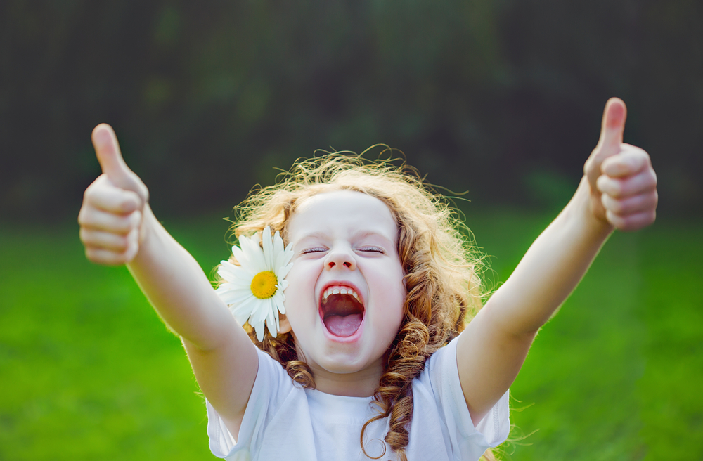 Joyful Young Girl Smiling with Thumbs Up Outdoors – Cosmetic Dentistry A cheerful young girl with curly hair and a daisy in her ear giving two thumbs up and smiling brightly in a green outdoor setting – Cosmetic Dentistry
