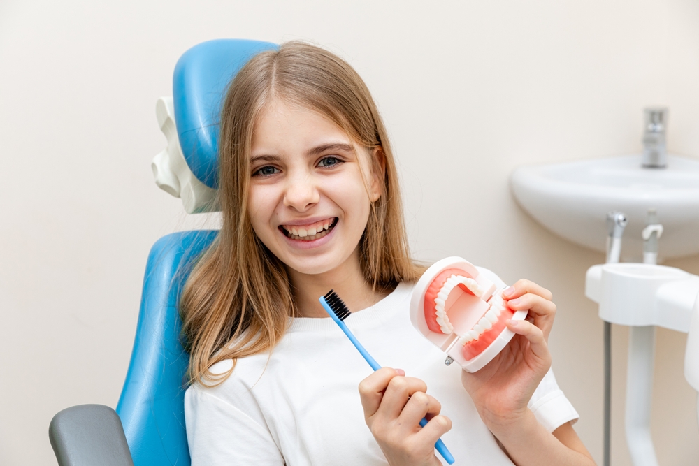 A smiling young girl in a dental chair holding a toothbrush and denture model, possibly before or after a tooth extraction consultation – Extractions