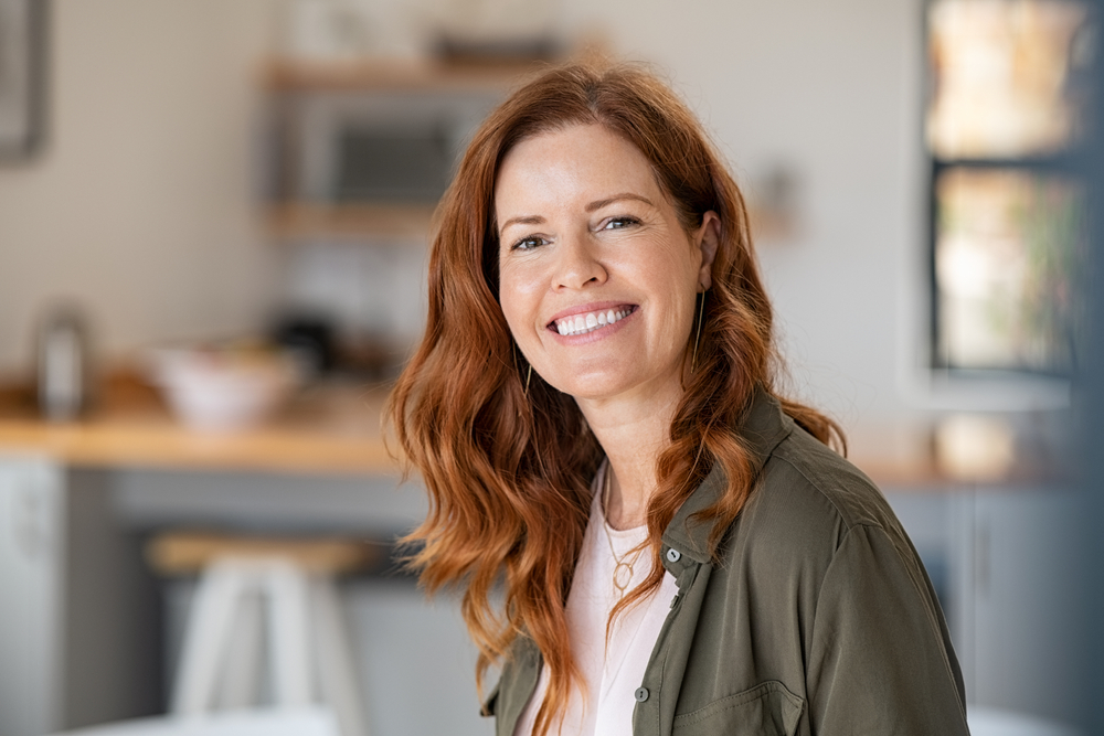A middle-aged woman smiling confidently in her kitchen, revealing clean and white teeth likely enhanced by professional whitening – Teeth Whitening
