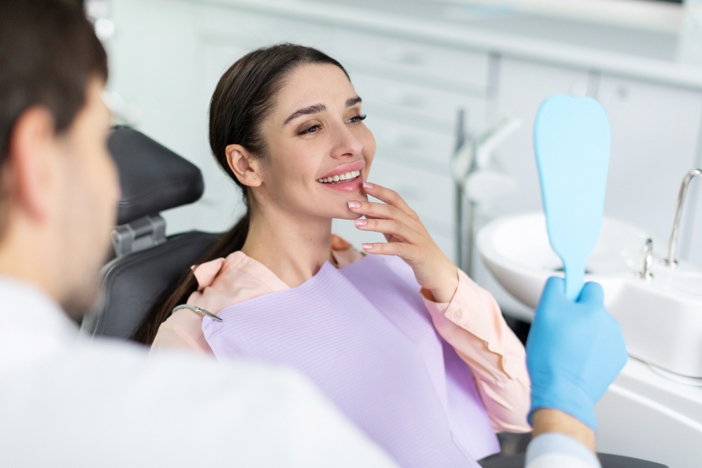 A woman sitting in a dental chair smiling while holding a mirror handed by a dentist - Dentist Graham WA A woman sitting in a dental chair smiling while holding a mirror handed by a dentist - Dentist Graham WA