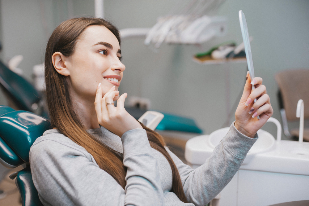Young Woman Admiring Smile After Dental Cleaning – Cleaning and Exams A woman in a dental chair smiling while looking at her reflection in a handheld mirror after a dental cleaning – Cleaning and Exams
