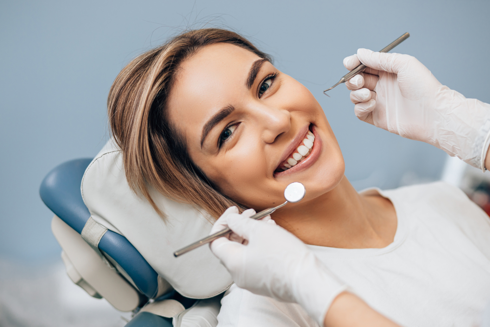 Smiling Woman During Dental Implant Checkup – Dental Implants A cheerful woman reclining in a dental chair while a dentist uses tools to examine her mouth, possibly during a dental implant follow-up – Dental Implants