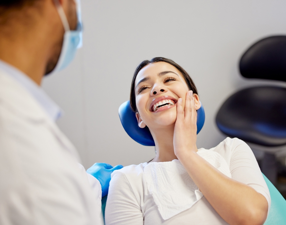 A young woman in a dental chair smiles and touches her cheek while speaking with a masked dentist, illustrating quality care and comfort – Dentist in South Hill WA