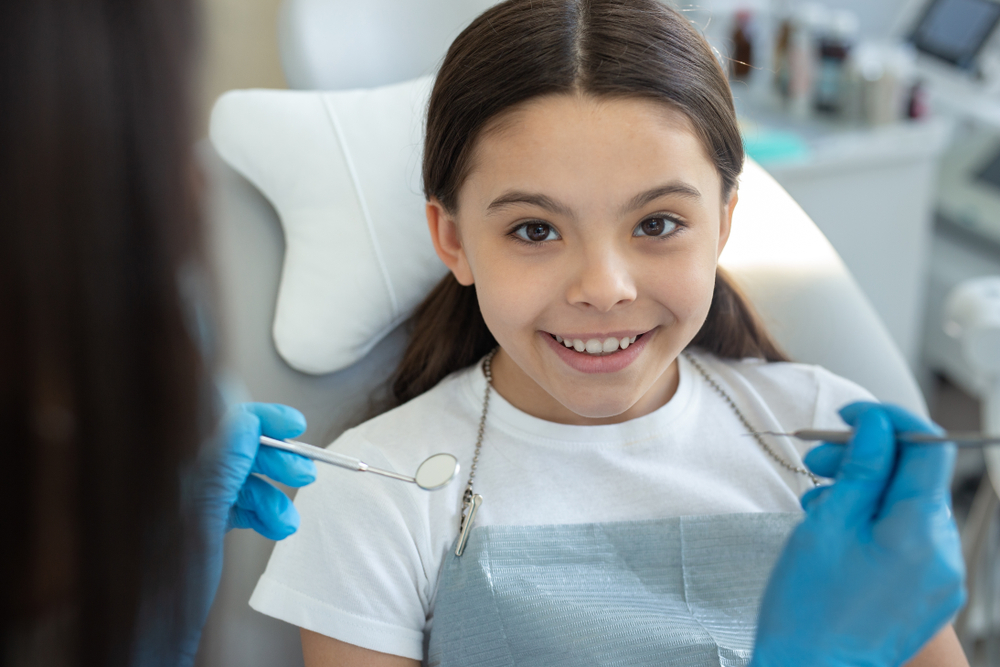 A cheerful young girl sitting in a dental chair, ready for her dental bonding procedure, as a dentist prepares with tools – Dental Bonding