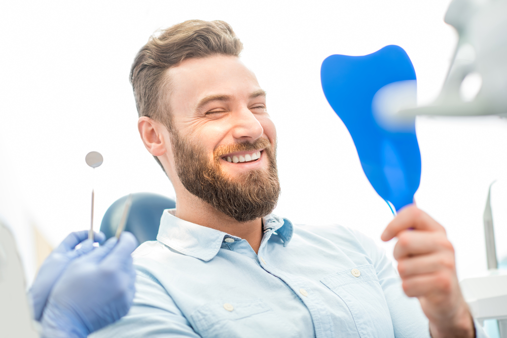 A cheerful man in a dental chair smiling while looking into a mirror, pleased with the results of his root canal or other dental treatment – Root Canals