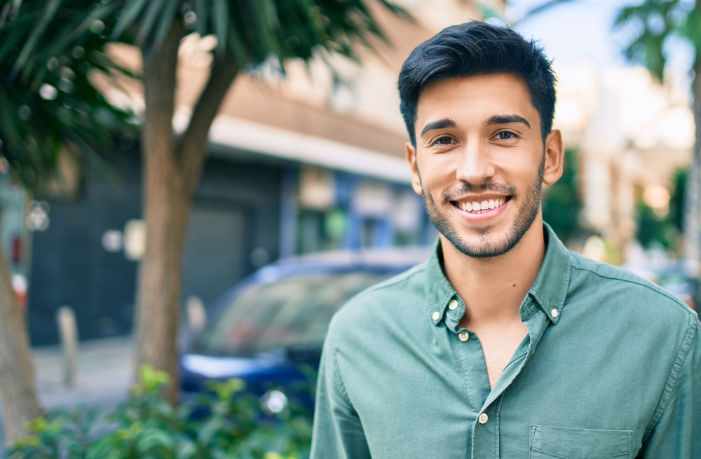 Confident Man Smiling Outdoors in Urban Setting – Dentist Orting WA A young man with a healthy, bright smile stands outdoors in a sunny neighborhood, showcasing the results of quality dental care – Dentist Orting WA