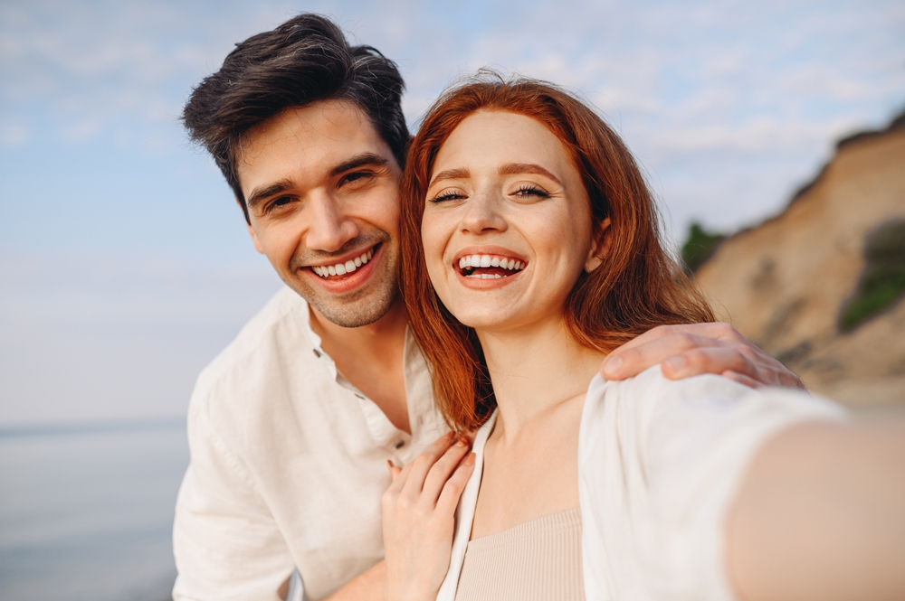 A joyful couple takes a selfie on a sunny beach, both smiling with bright, healthy teeth, reflecting confidence in their dental care – Dentist Spanaway WA