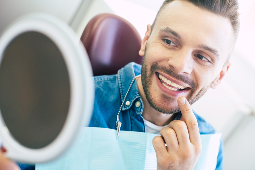 Smiling Man Examining Dental Work in Mirror – Dental Implants A happy man sitting in a dental chair, pointing to his teeth while looking in a handheld mirror, checking the results of his dental implants – Dental Implants